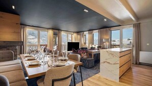 A modern dining area with a long table set for six, a kitchen area with a marble island, and large windows offering a view of the mountains. - Luxurious Apartment with Ski-In and Out Access in Centre (Val Thorens)
