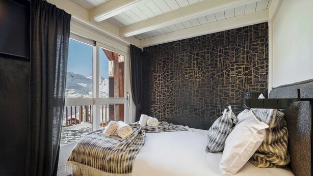 A hotel room with a bed, a window with curtains, a patterned wall, and a view of mountains. - Luxurious Apartment with Ski-In and Out Access in Centre (Val Thorens)