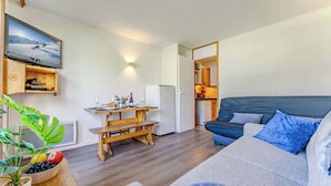 Ein gemütliches Wohnzimmer mit einer hölzernen Bank, einem Sofa mit blauen Kissen, einem Esstisch mit Flasche und Gläsern sowie einer durchgehenden Tür erkennbaren Küche.