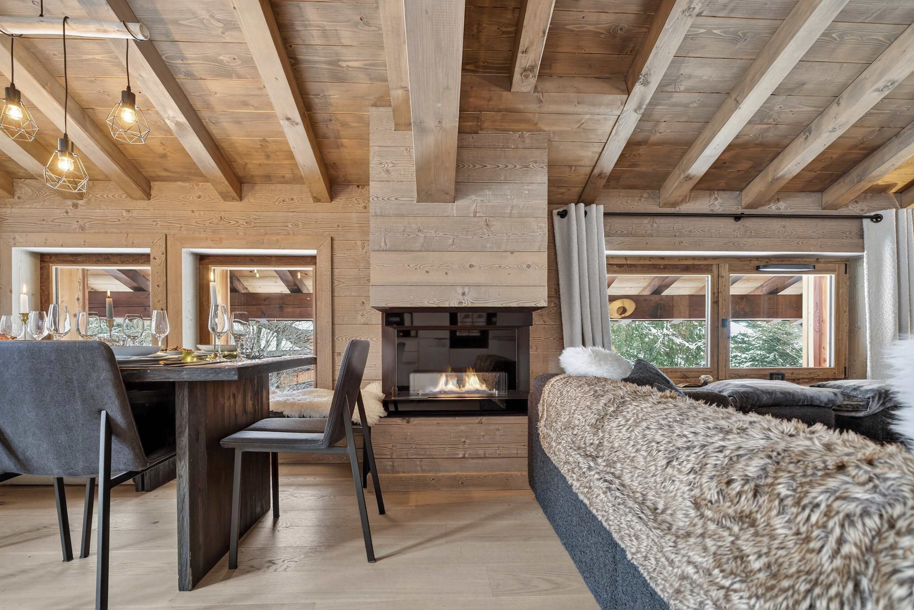 A cozy room with a fireplace, wooden beams, a dining area with chairs and a table set for two, and large windows offering a view of the outdoors.