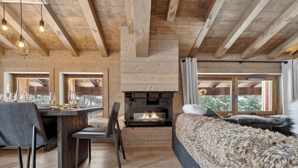 Ein gemütlicher Raum mit Kamin, Holzbalken, einer Essecke mit Stühlen und einem Tisch für zwei Personen sowie großen Fenstern mit Blick ins Freie.