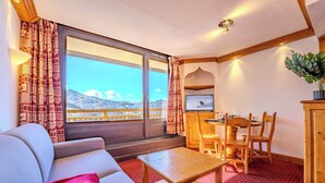 A cozy room with a sofa, a wooden table, a dining area with chairs, and a view of mountains through large windows. - Alpine Stay w/ Private Ski Views (Val Thorens)