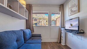 Um espaço compacto com um sofá azul, uma televisão sobre um suporte, e vista para edifícios por uma porta.
