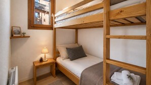 Habitación con literas, ventana, mesita de noche, lámpara y una pequeña estantería con un cuadro.