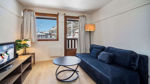 Una sala de estar moderna con un sofá azul oscuro, una mesita redonda pequeña, un televisor sobre un mueble de madera, una lámpara y vistas a una montaña nevada a través de una ventana.