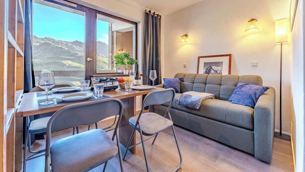 Quarto aconchegante com uma mesa de jantar posta para dois, um sofá e vista para montanhas através de grandes janelas.