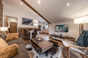 Ein modernes Wohnzimmer mit einer Couch, einem Couchtisch und einem Sessel. Im Hintergrund befindet sich eine Küche mit Schränken und einem Essbereich.