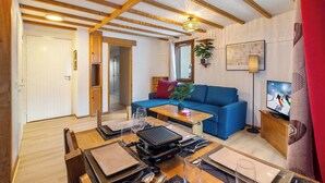 A modern living room with a blue sofa, wooden ceiling beams, a dining table set for four, and a television on a wooden stand. - Ski-In and Out Mountain View Escape 47 (Val Thorens)