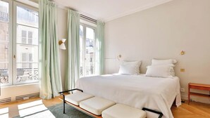 A bedroom with a large bed, a bench, a window with curtains, and a view of buildings outside. - EXCEPTIONAL SETTING ON RUE BUCI TO EXPERIENCE PARIS AT FINEST, SAINT-GERMAIN (Paris)