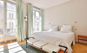 Ein Schlafzimmer mit einem großen Bett, einer Bank, einem Fenster mit Vorhängen und Blick auf Gebäude.