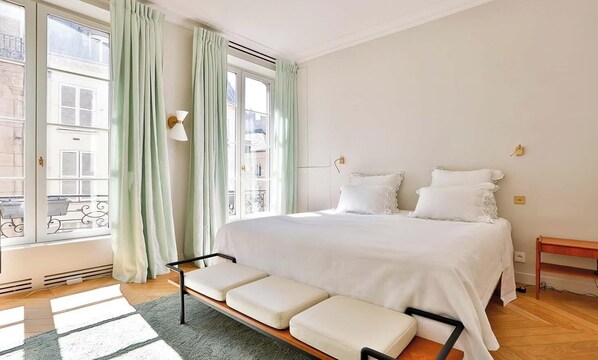 Quarto com cama grande, um banco, janela com cortinas e vista para prédios.