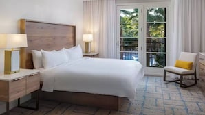Camera d'albergo con un letto grande, due comodini con lampade, una sedia e vista su palme attraverso una porta vetrata.