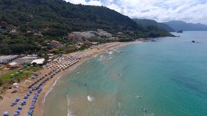 A beach with rows of umbrellas and beach chairs, a sandy shore, and buildings in the background. - Villa Triton (Corfu)