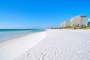 Ein Strand mit weißem Sand, klarem, blauem Wasser und einer Reihe mehrstöckiger Gebäude im Hintergrund.