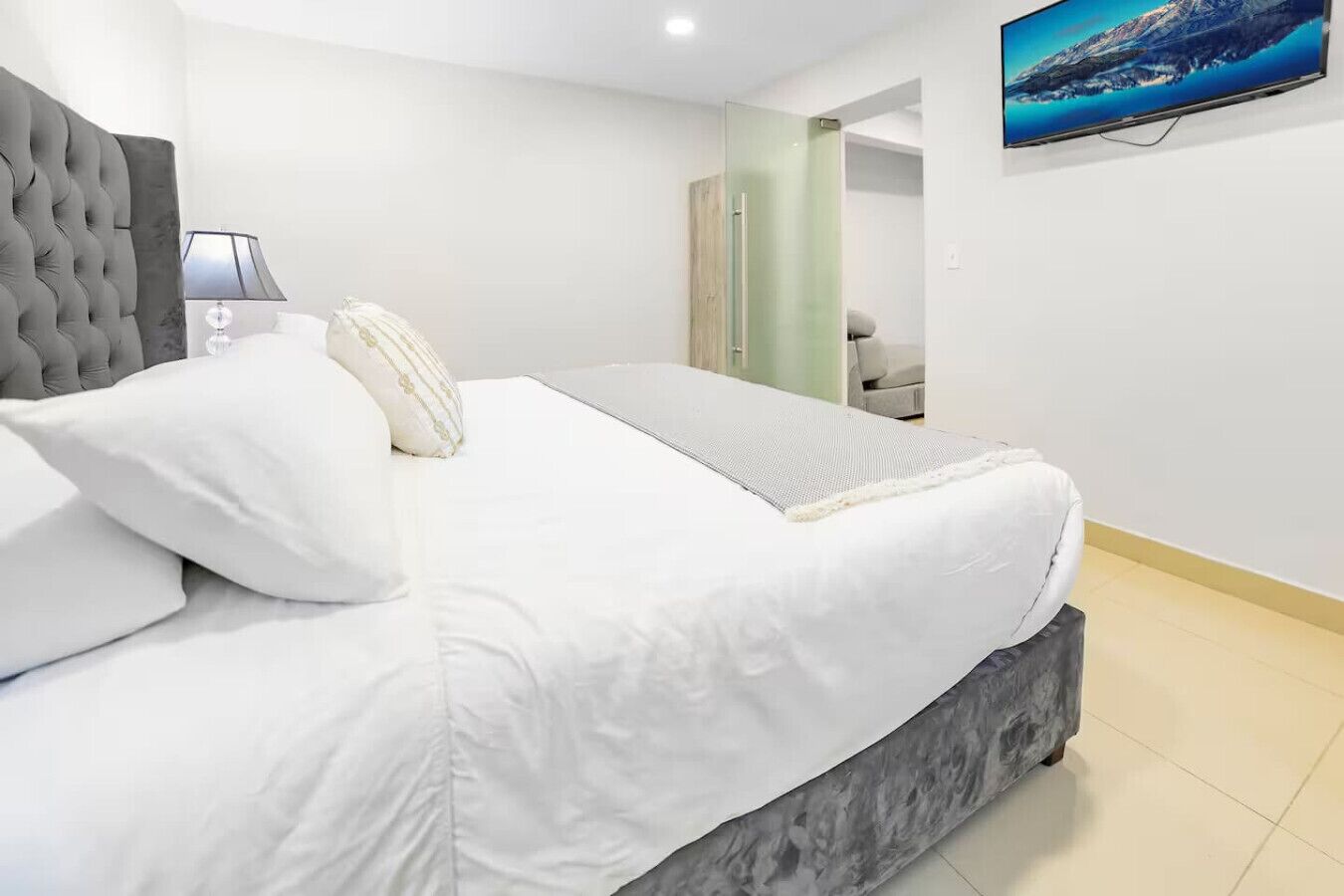 Een moderne slaapkamer met een groot bed, een grijs gestoffeerd hoofdeinde, een bedlampje en een flatscreen televisie aan de muur.
