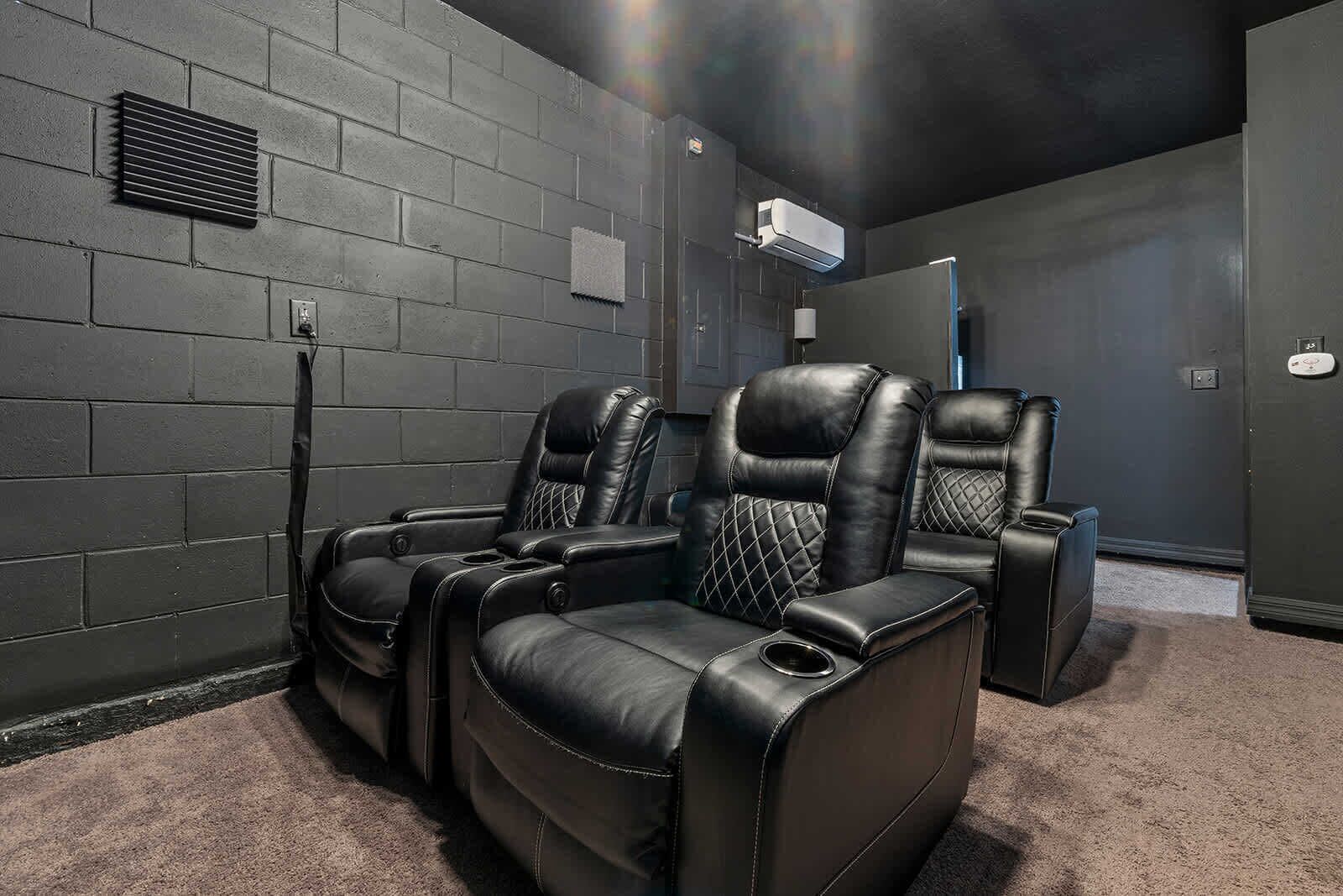 Une salle de cinéma maison avec trois fauteuils inclinables en cuir noir, un sol recouvert de moquette et un mur en briques sombres.