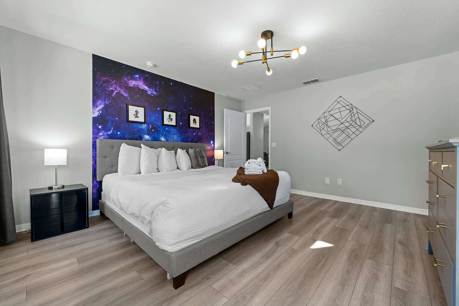 A modern bedroom with a galaxy-themed wall mural, a grey bed, a bedside table with a lamp, and a geometric wall art piece.