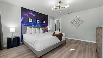 A modern bedroom with a galaxy-themed wall mural, a grey bed, a bedside table with a lamp, and a geometric wall art piece.