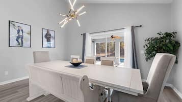 Un coin repas avec une table blanche, des chaises et un lustre moderne. Deux tableaux encadrés ornent le mur, ainsi qu’une plante. On peut également admirer le paysage extérieur à travers la fenêtre.