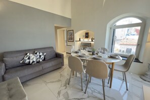 A modern living room with a grey sofa, dining table set for four, and a large window with a view of buildings. - Exclusivity, Luxury and Sea View with 2 Bedrooms (Alassio)