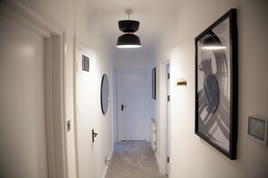 A hallway with white walls, a black-framed abstract painting, a ceiling light, and a door at the end. - Luxury 2 BED near tube station (London)