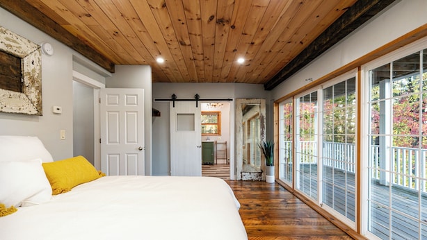 A bedroom with a wooden ceiling, a large bed with white and yellow bedding, and a view of a balcony with wooden railings. - Lockes Hill Retreat - Close to Gunstock (Gilford)