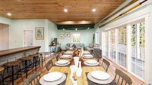 A dining area with a wooden table set for six, surrounded by bar stools, and a view of the outdoors through large windows. - Lockes Hill Retreat - Close to Gunstock (Gilford)