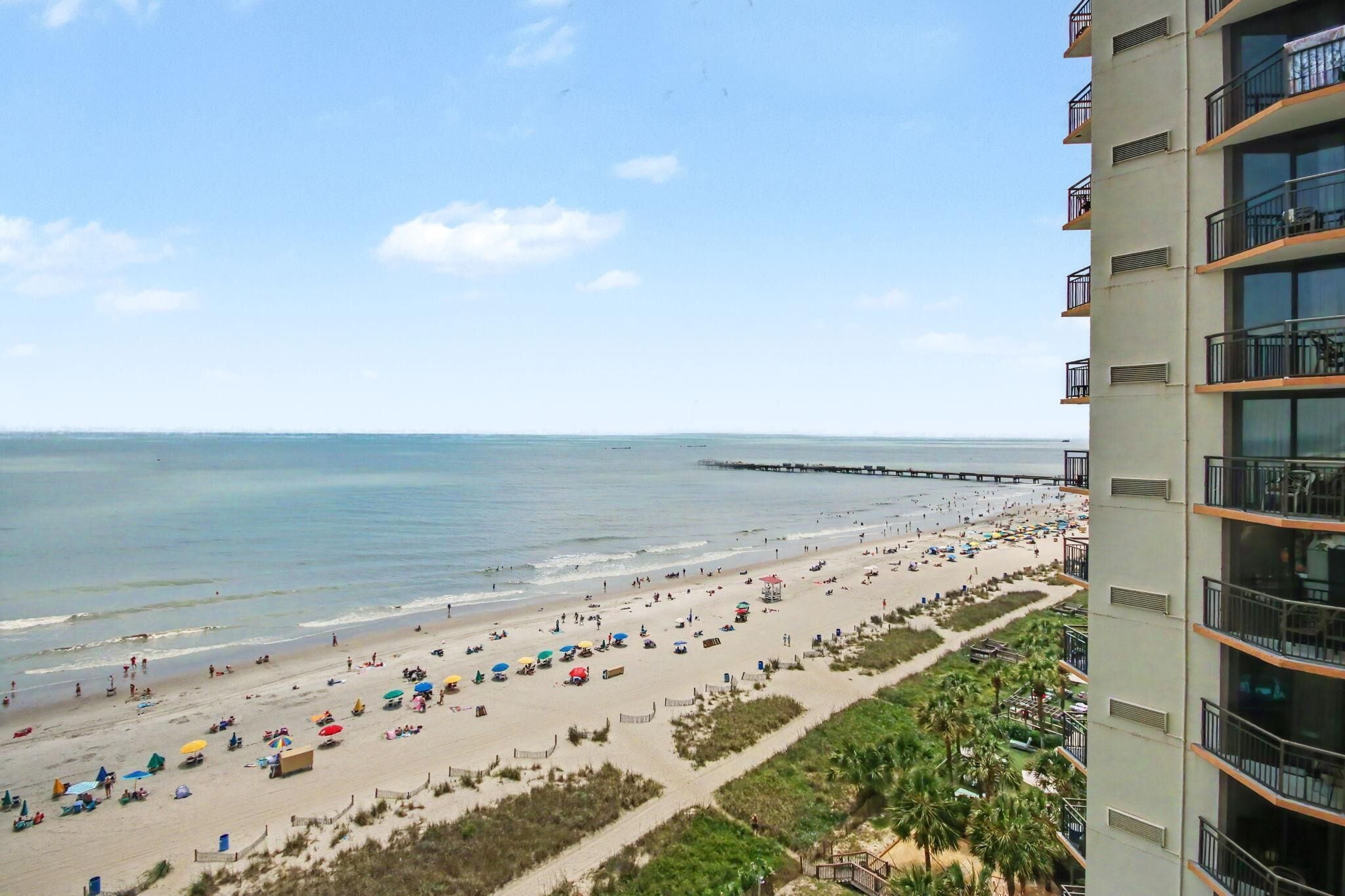 A beach with umbrellas and people, a pier, and a building with balconies.