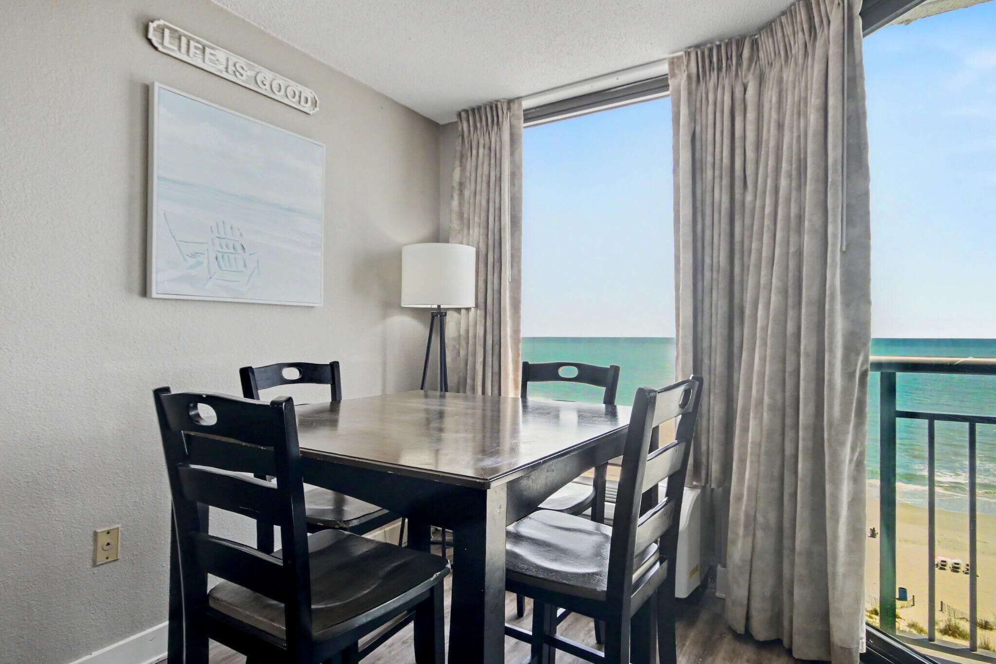 A dining area with a table and chairs, a lamp, a framed picture, and a view of the beach.