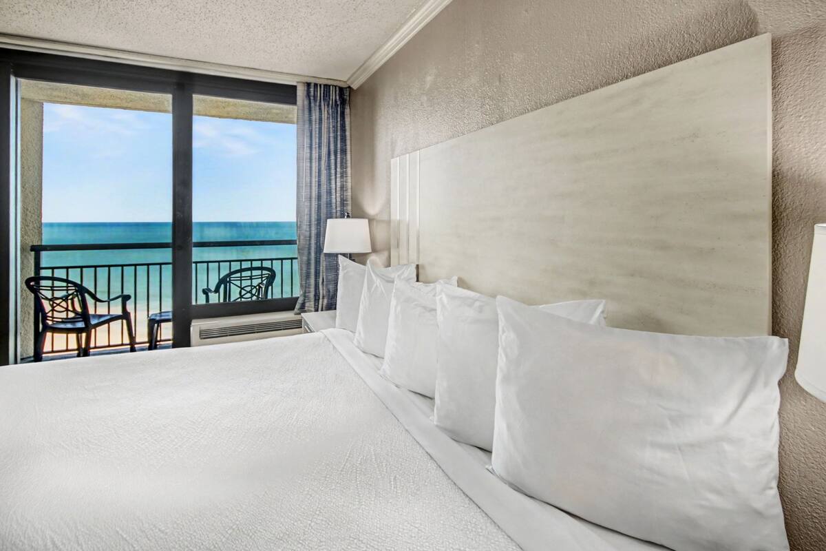 Ein Hotelzimmer mit einem großen Bett, Meerblick und einem Balkon mit Sitzgelegenheiten.