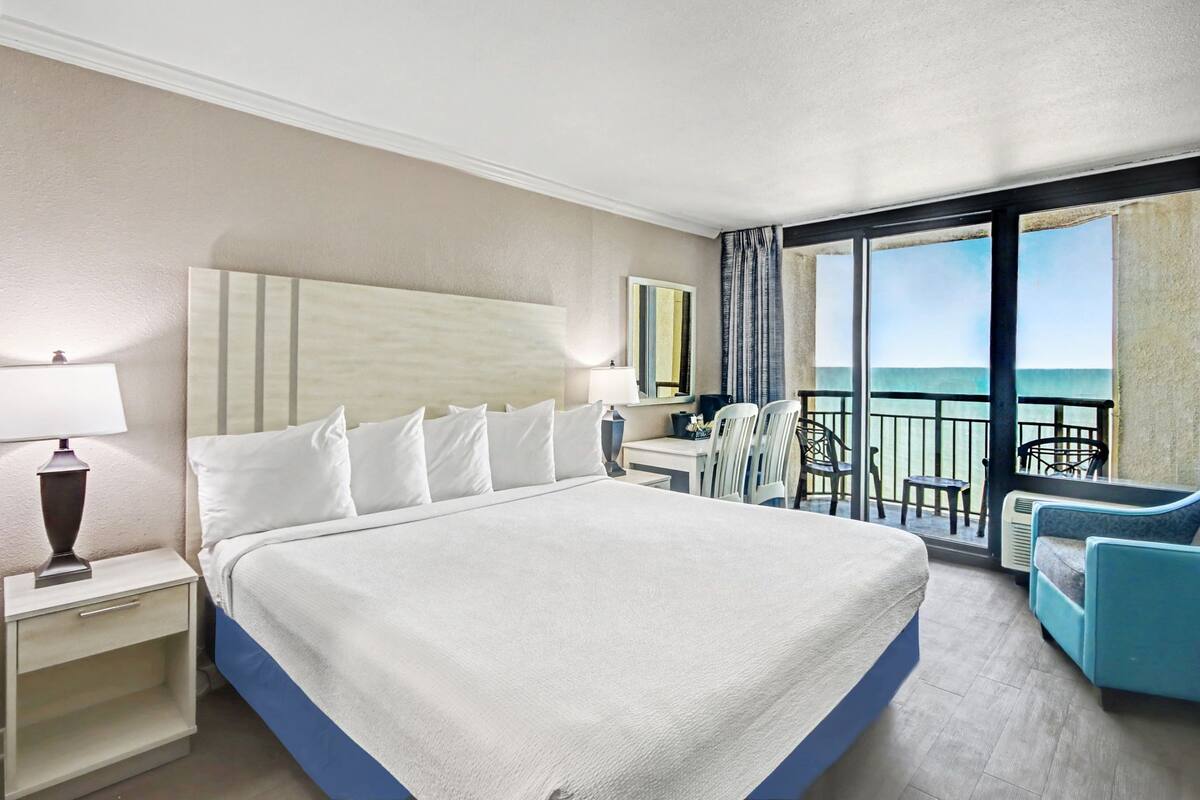 Ein Hotelzimmer mit einem großen Bett, einem Schreibtisch, einem Stuhl und einem Balkon mit Meerblick.