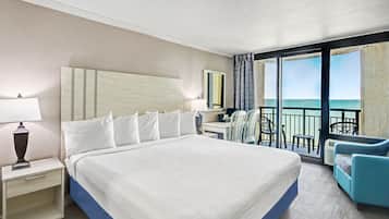 A hotel room with a large bed, a desk, a chair, and a balcony with a view of the ocean.