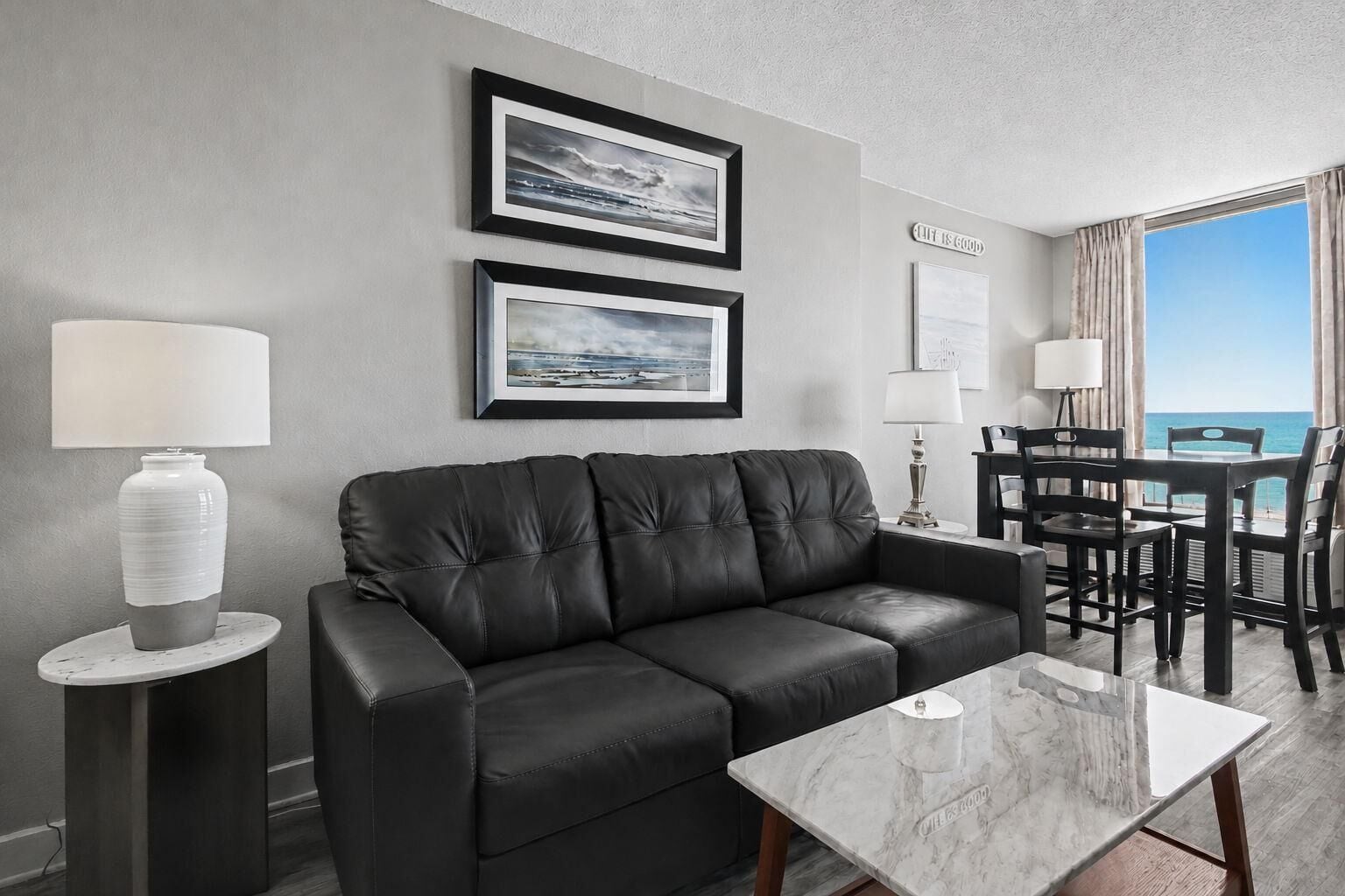 A modern living room with a black leather sofa, two framed pictures, a dining table with chairs, and a view of the ocean through large windows.