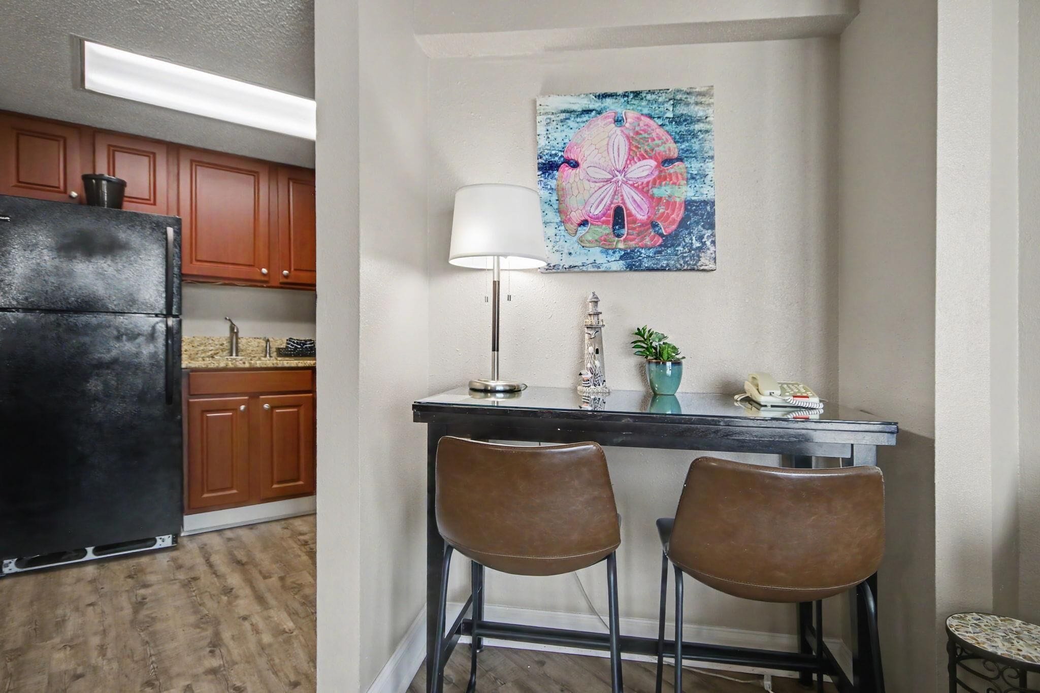 Una cocina con armarios de madera, un refrigerador negro, una mesa de comedor con dos sillas, una lámpara y un cuadro decorativo.