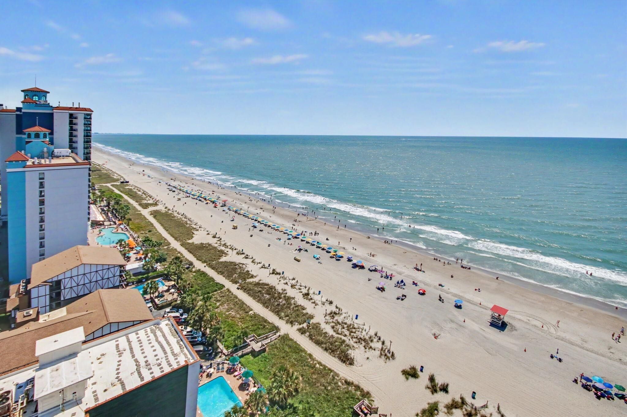 A beach resort with a long sandy beach, a pool, and a hotel building with multiple floors and balconies.