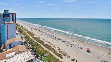 A beach resort with a long sandy beach, a pool, and a hotel building with multiple floors and balconies.