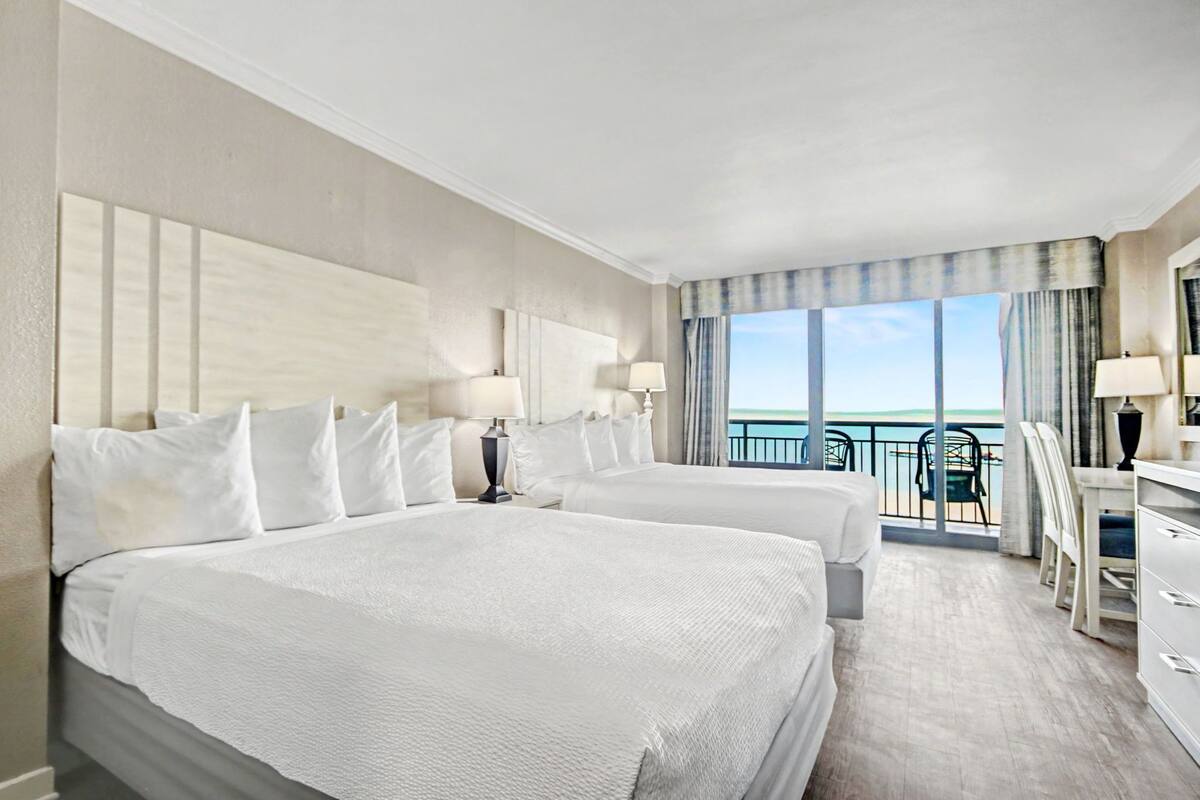 Ein Hotelzimmer mit zwei Betten, einem Balkon mit Meerblick und einem Schreibtisch mit Stuhl.