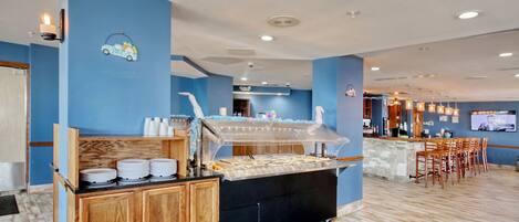 Un'area buffet con un bancone, piatti e bicchieri, e una zona bar con sedie e uno schermo televisivo.