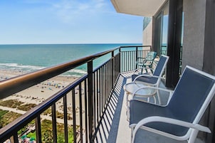 A balcony with a view of the beach and ocean, equipped with chairs and a small table.