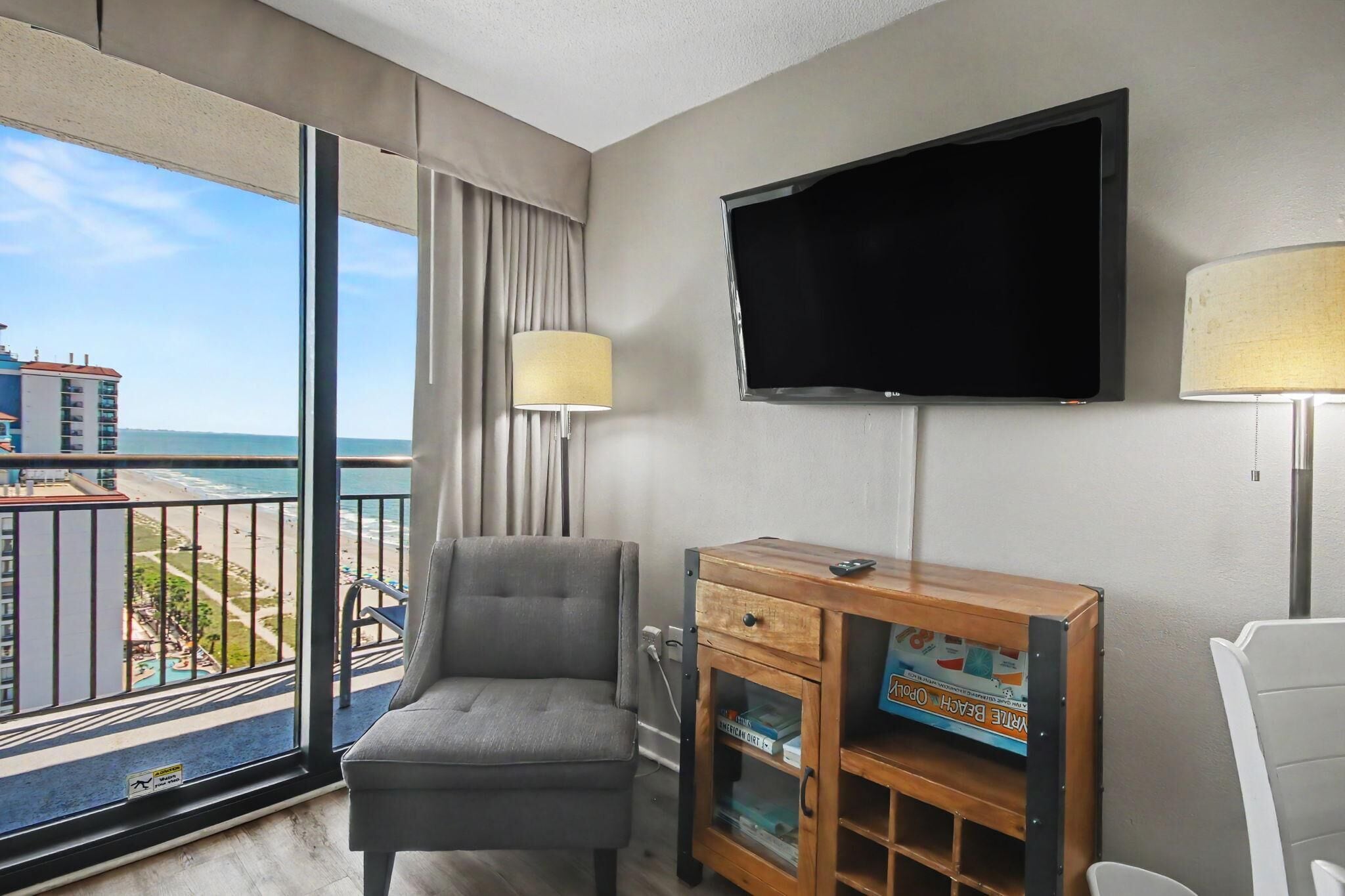 A hotel room with a large window, a flat-screen TV, a grey armchair, a wooden side table, and a lamp.
