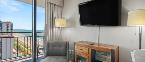 A hotel room with a large window, a flat-screen TV, a grey armchair, a wooden side table, and a lamp.