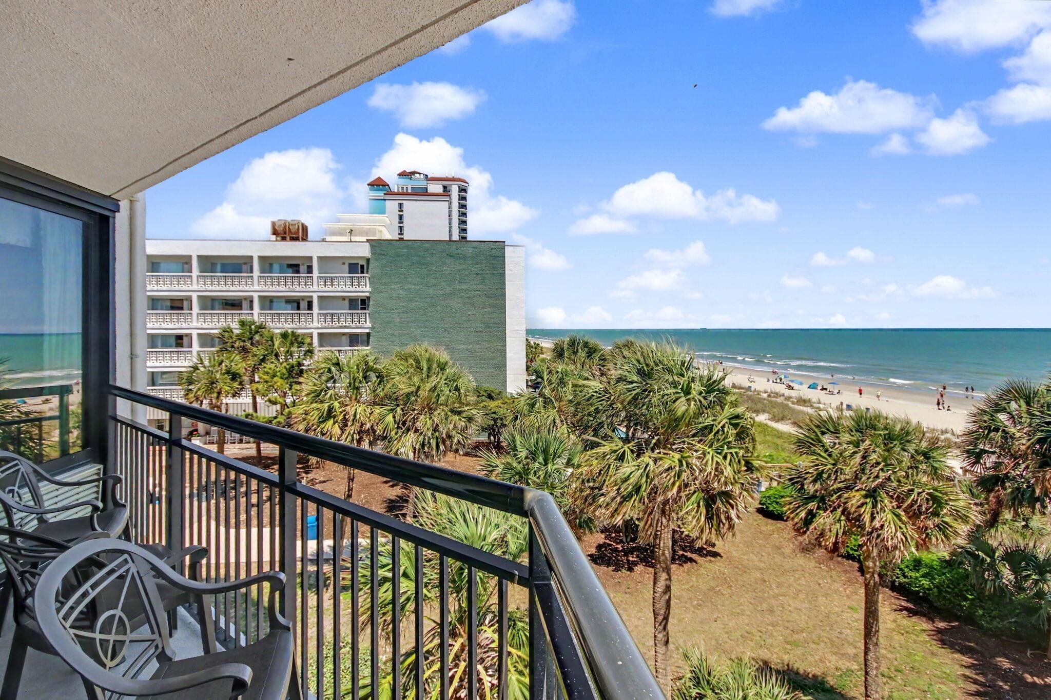 A balcony with a view of a beach, palm trees, and a hotel building in the background.