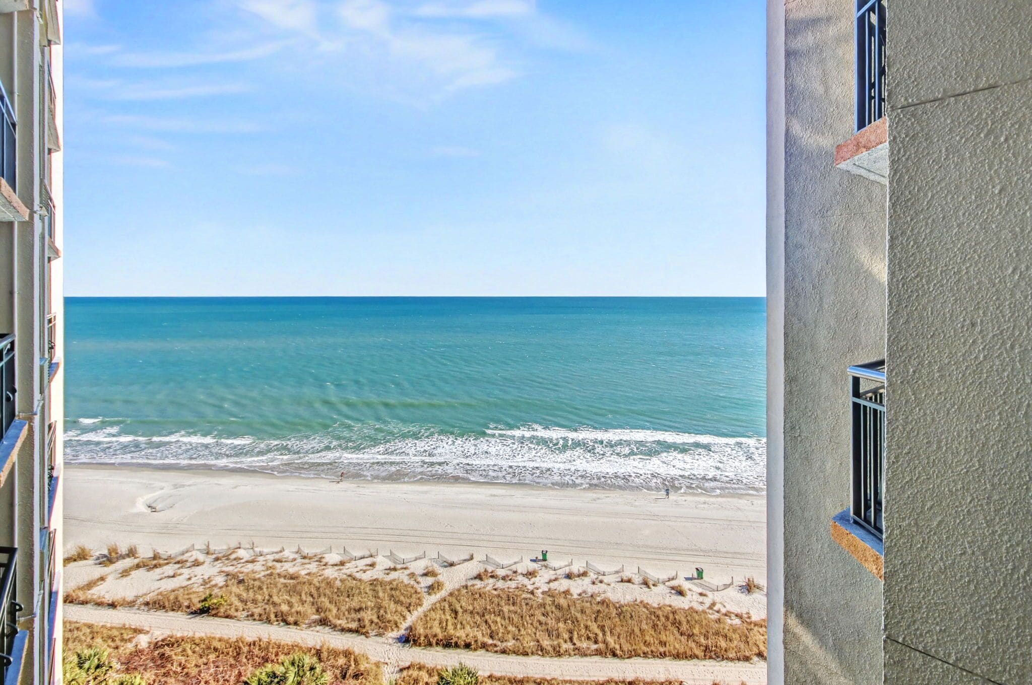 A view of a beach from a building with a balcony, featuring a sandy beach, ocean, and clear sky.