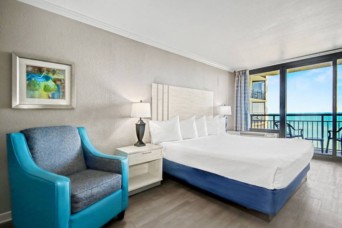 Ein Hotelzimmer mit einem großen Bett, einem Nachttisch mit Lampe, einem blauen Sessel und einem Balkon mit Meerblick.