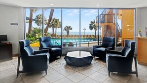 Un hall de hotel con un ventanal grande que ofrece vistas a una piscina y palmeras. Hay cuatro sillones azul oscuro y una mesa de centro hexagonal.