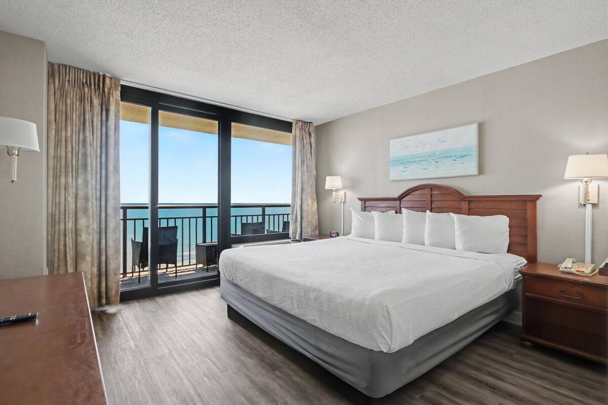 Ein Hotelzimmer mit einem großen Bett, einem Schreibtisch, einem Stuhl, einem Nachttisch, einer Lampe, einem Gemälde und Blick aufs Meer.