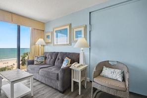 A room with a sofa, a small table, a lamp, a wicker chair, and framed pictures on the wall.