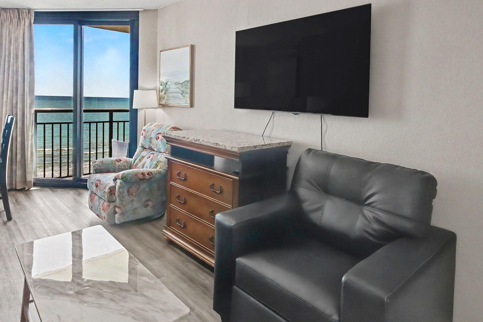 Un soggiorno con un divano in pelle nera, un settimino in legno, una televisione montata a parete e un'ampia finestra vista mare.