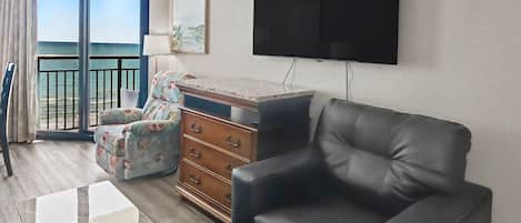 Un soggiorno con un divano in pelle nera, un settimino in legno, una televisione montata a parete e un'ampia finestra vista mare.