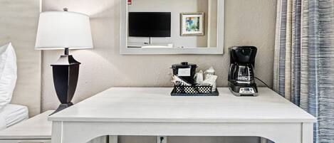 Een hotelkamer met een wit bureau, een zwarte lamp, een koffiezetapparaat, een ingelijst beeld en een televisie.
